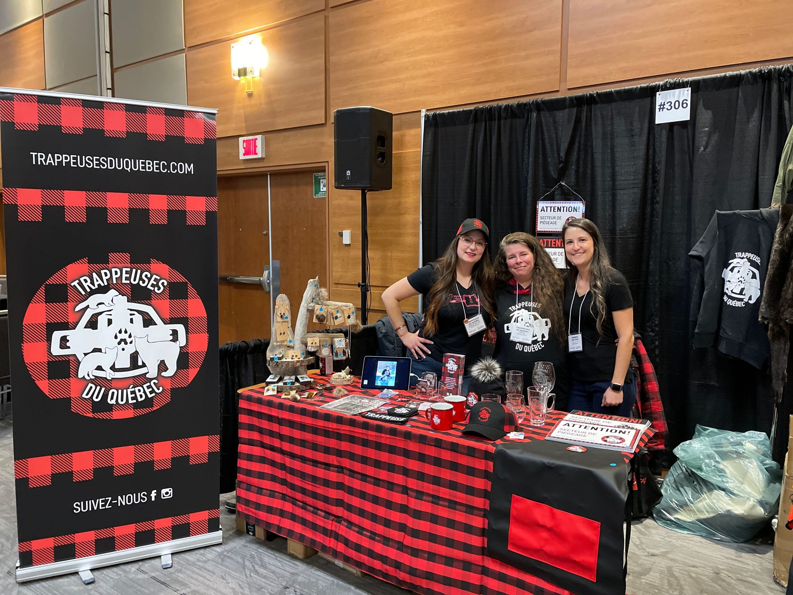 Stéphanie, à gauche de deux accolytes, sont au kiosque des Trappeuses du Québec. Devant elle, une table recouverte d'une nappe carreautée rouge et noire. Plusieurs items promotionnels se trouve sur cette table. À leur gauche, un grand parapost avec le logo des Trappeuses du Québec. L'affiche est aux couleurs de la nappe, soit carreautée rouge et noire. 